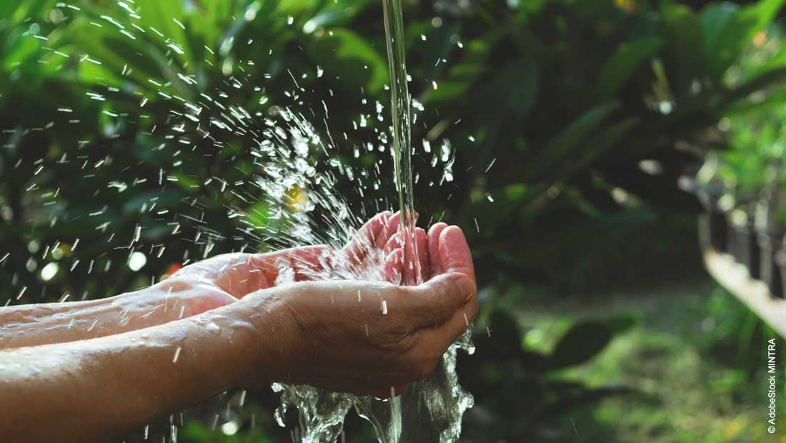 Ein Bild einer Hand, die Wasser aus einer Quelle in eine offene Handfläche gießt. Im Hintergrund ist eine satte grüne Landschaft zu sehen.
